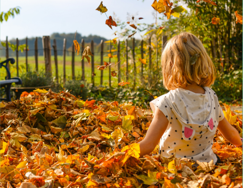 A young girl's back, kneeling amongst a garden of autumn leaves and gazing through a fence into a field of greenery with trees in the distance. The young girl is wearing a white and black polka-dot dress. #nature #autumn # leaves #fluseason #fluvaccination