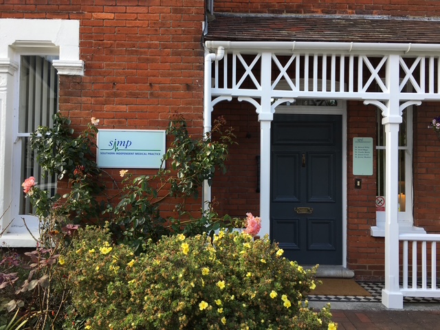 Front entrance of the Southern Independent Medical Practice, with a dark blue door, white wooden trim, brick facade, and flowering plants in the foreground.