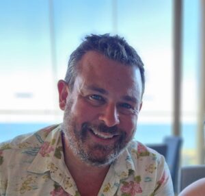 A man smiling indoors with a soft ocean view in the background.
