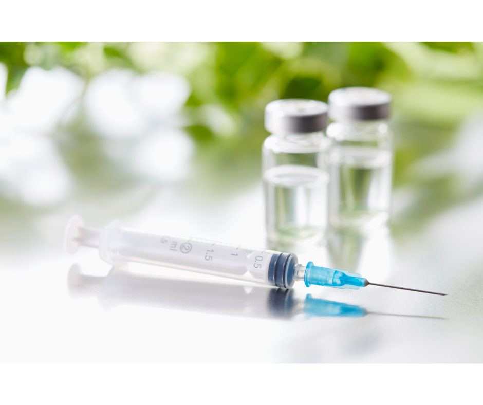 A syringe with a blue needle lying on a reflective surface, with two clear vaccine vials in the background.